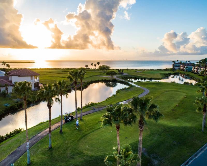 An aerial view of a bright green golf course over looking the bay as the sun shines on the water