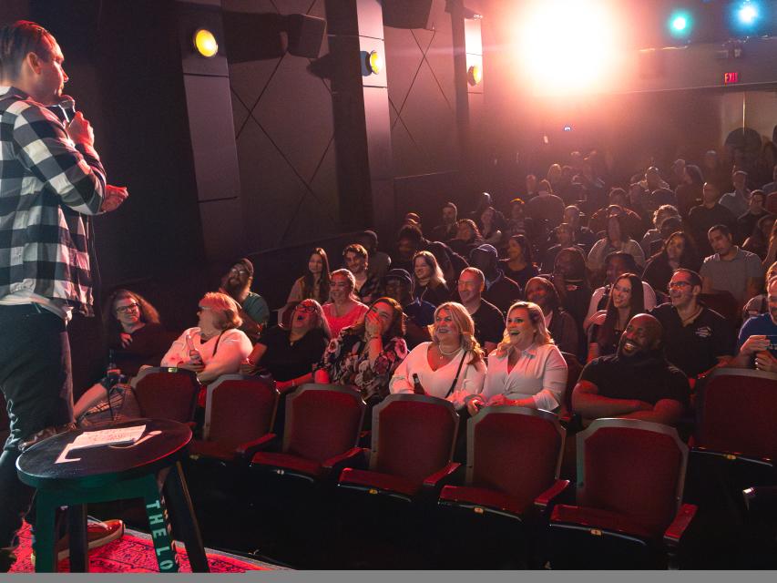Stand-up performer on stage facing a packed theater audience, who are laughing under bright stage lights.