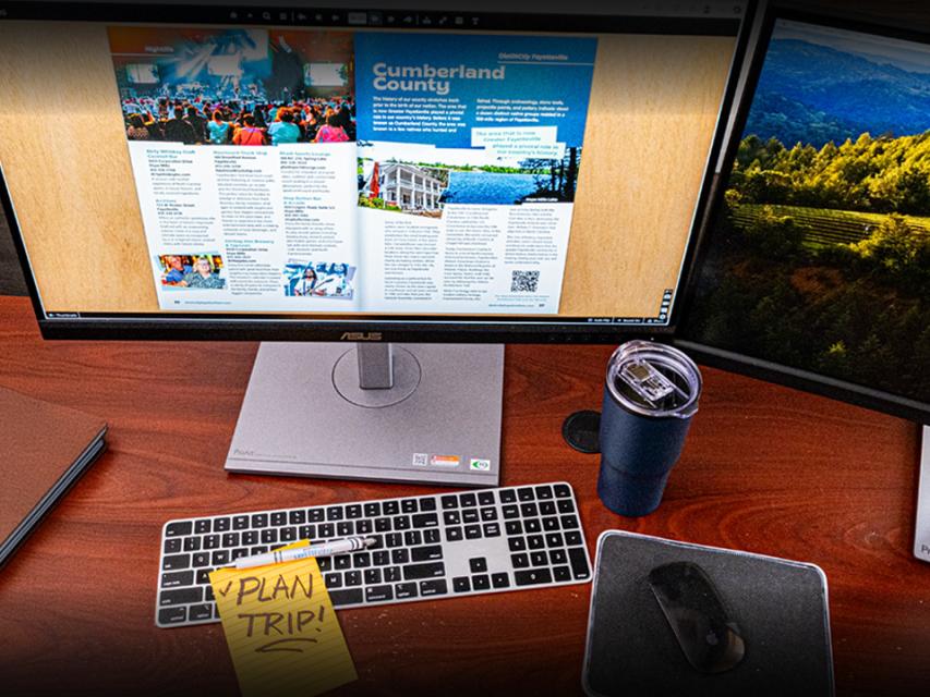 A digital destination guide is open on a computer monitor at an office desk with a keyboard, mousepad, a blue travel cup, and a yellow sticky note that says Plan Trip.