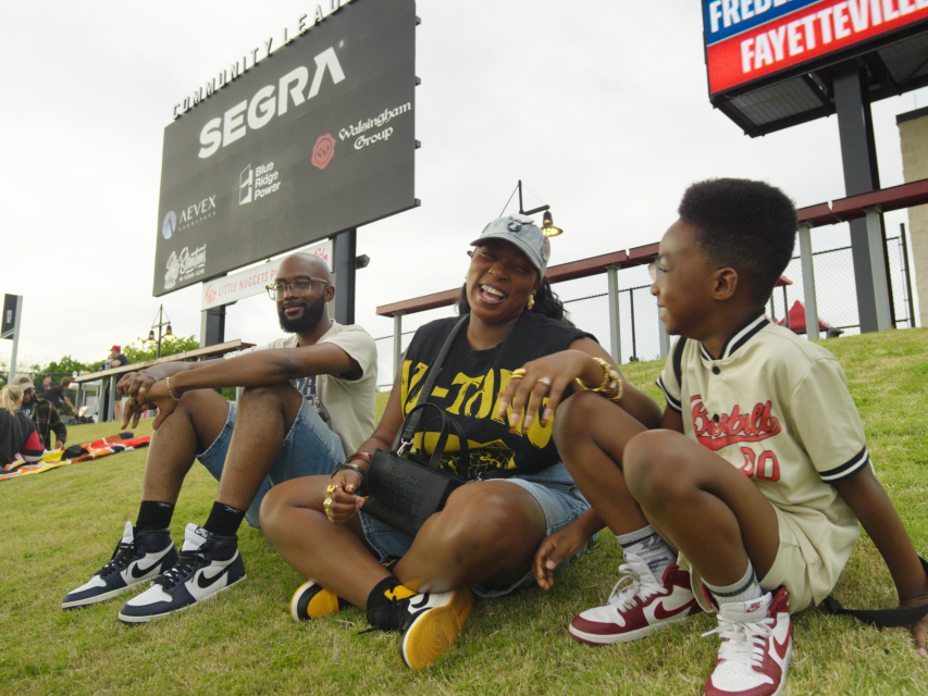 Family sitting on a grassy hill at Segra Stadium in Fayetteville, smiling and talking during a baseball game