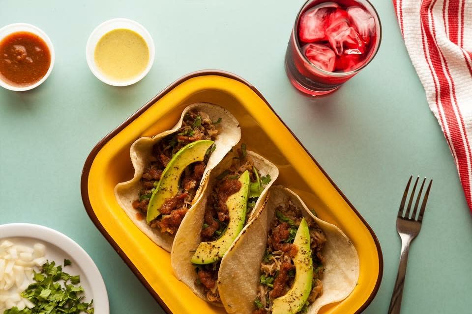Image of a yellow plate with three tacos sitting on a table with two sides of salsa and a beverage.