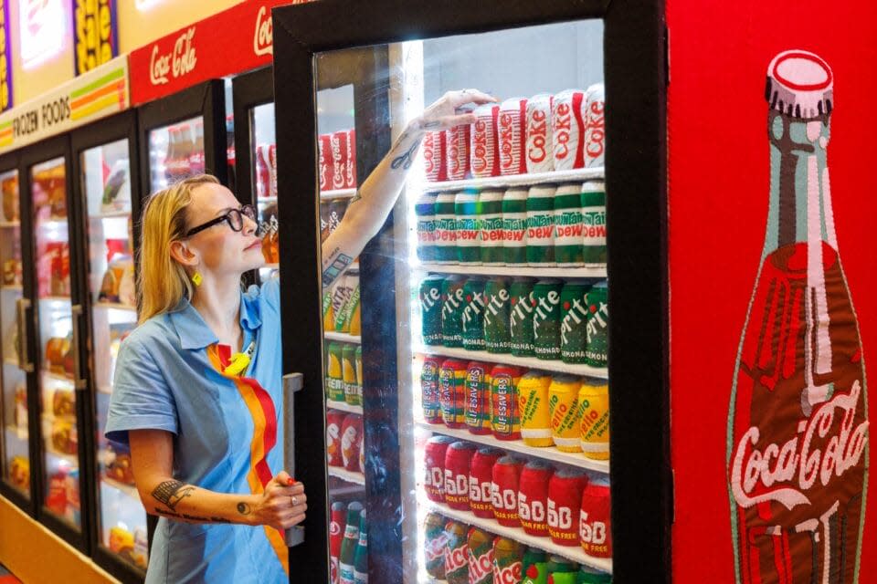 Lucy Sparrow opens the glass door of a refrigerated beverage case inside Lucy Sparrow's The Beginning of Convenience installation at the Momentary, reaching toward rows of hand-stitched felt soft drink cans, including Coca-Cola, Diet Coke, Sprite, Mountain Dew, and other colorful beverages displayed like a convenience store cooler.