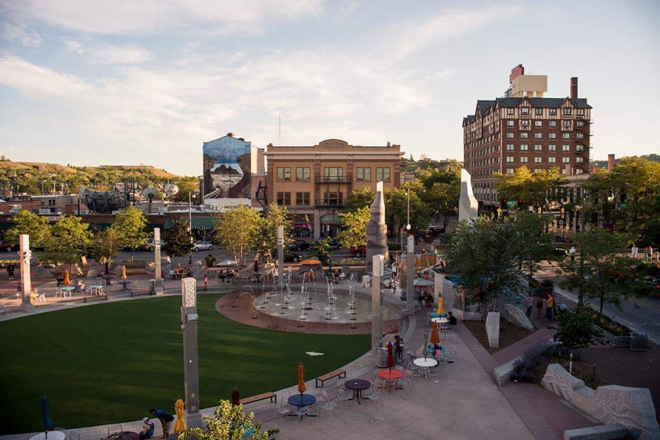 View of patch of green space with a splash pad surrounded by historic buildings in a charming downtown setting.