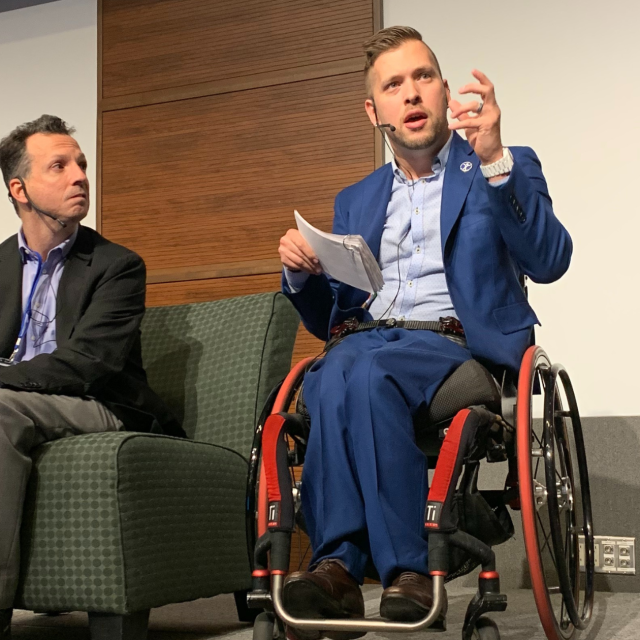Two men are on stage, one holding notes and speaking to the audience from a wheelchair, the other man listening from a chair.
