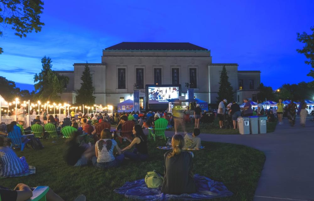 A crowd of people sit down on grass and watch a movie on a large screen outdoors. Behind the screen is a large building.