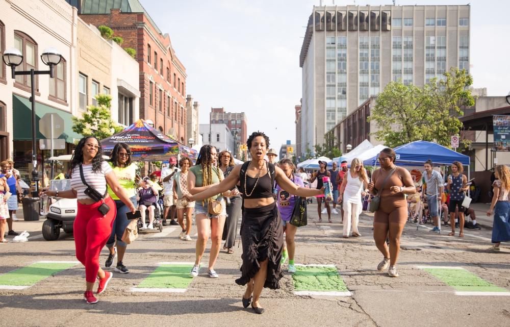 Women dance in the street during a major festival. Behind the dancing women, there are vendor booths that line the street.