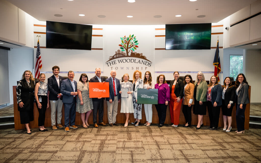 Seventeen people line up in front of dais in The Woodlands Township Board Chambers. They are all Visit The Woodlands Board Members and Staff. From left to right: Josie Lewis (Director of Sales), Brooklyn Newell (Social Media Coordinator), Ryan Greenwood (Senior Sales Associate), Nick Wolda (President), Cameron Klepac (Secretary / Treasurer of the Board), Brad Bailey (Director of the Board), Richard Franks (Vice Chairman of the Board), Linda Nelson (Chairman of the Board), Kim Lowe (Director of the Board), Noemi Gonzalez (Director of the Board), Monique Sharp (Director of the Board), Elizabeth Eddins (Executive Director), Carla Twiddel (Administrative Analyst), Julie Quinn (Servicing Specialist), Amber Parsley (Tourism Specialist), Ashley White (Director of Marketing), Andrea Day (Creative Media Coordinator). Missing: Ashley Fenner (Senior Sales Associate) and Rachael Varner (Content Coordinator)