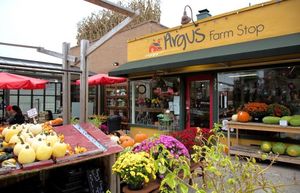 Farm stand storefront with produce, flowers, and pumpkins displayed out front. "Argus Farm Stop" is painted on the building.