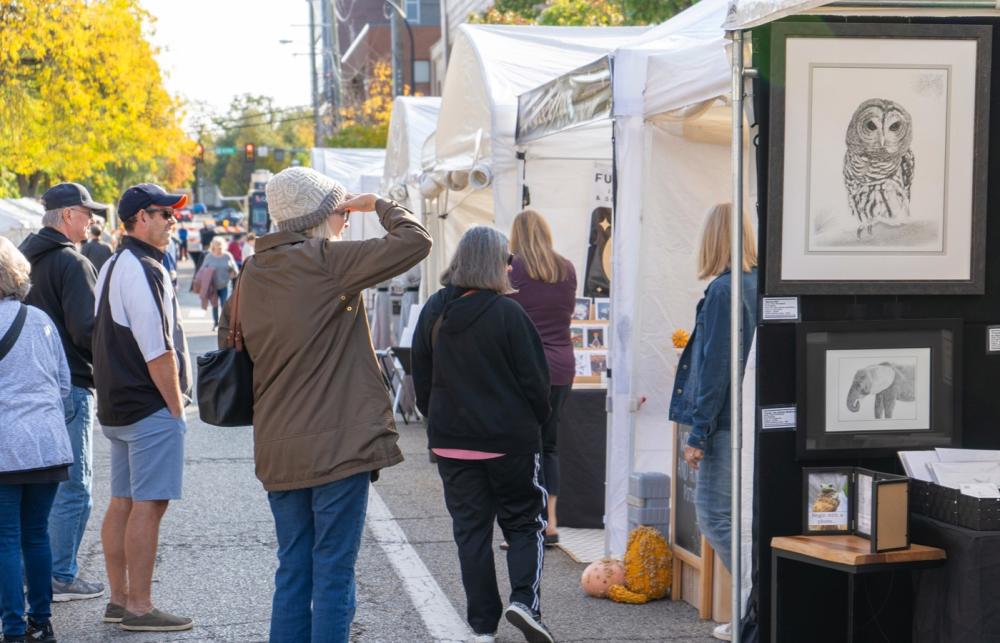 Woman stands with her hand shading her eyes as she admires artwork hanging in a vendor booth during a street art fair. People to the back and sides also viewing nearby artwork.