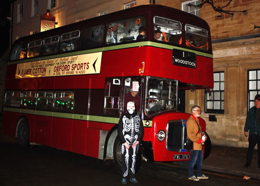 Vintage red double-decker bus from the Oxford Bus Museum in Woodstock in the Cotswolds, illuminated at night with visitors in Halloween fancy dress nearby.