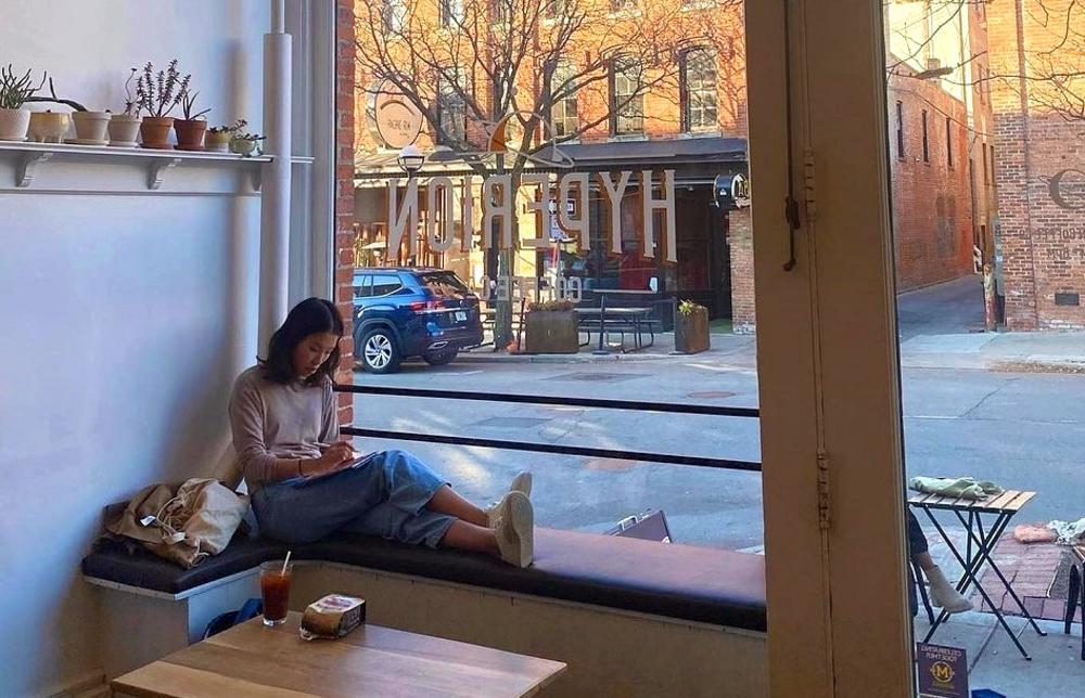 Woman sits on a ledge inside  a coffee shop, writing in a notebook.