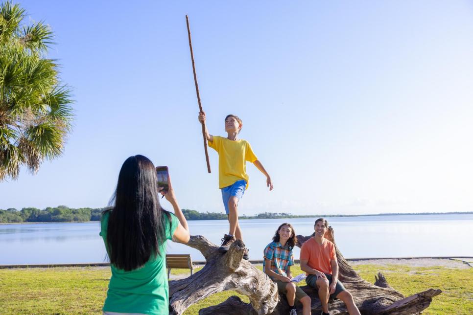 A boy stands on a tree limb while his mother takes a photo.