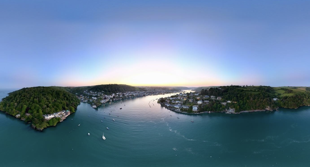 Aerial view of a tranquil river winding between two lush, green hills, dotted with small boats. Houses line the riverbanks under a clear, pastel sunset sky.