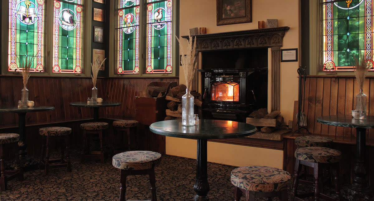 Cozy pub seating area at County Clare Irish Pub with small round tables, wooden stools, stained glass windows and a glowing fireplace.