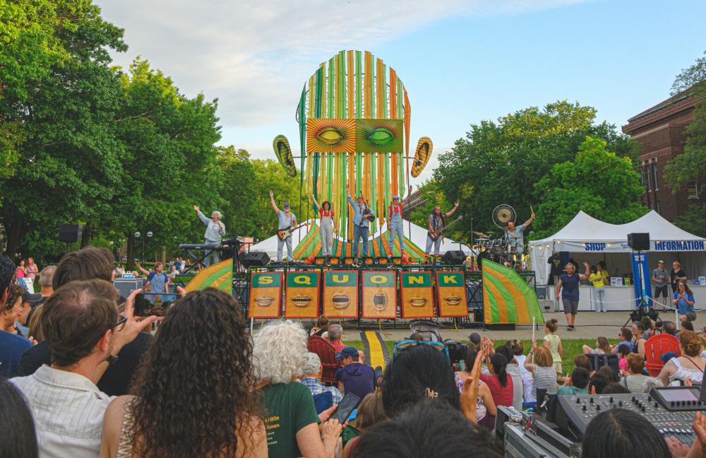 Band of 7 bandmates stand and wave to the crowd as they stand on a custom stage with a background shaped like a man's head with ears.  The stage read reads "SQUONK" below the bandmates' feet.