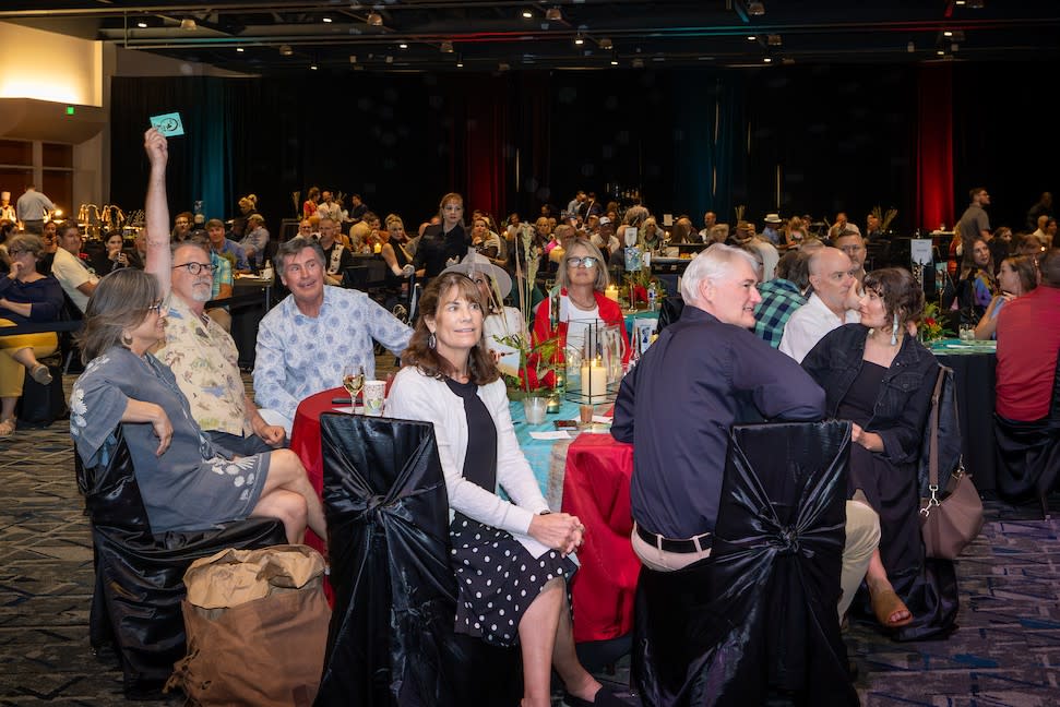 The camera overlooks a ballroom at The Woodlands Waterway Marriott. There are a dozen or more round tables with classy decor and catered food. The guests are all adult men and women, each dressed as if for different events (casual to semi-formal). Everyone looks to the stage off-screen to the right, presumably to bid on art pieces for sale. One man on the left raises a blue card in the air to signify his bid.