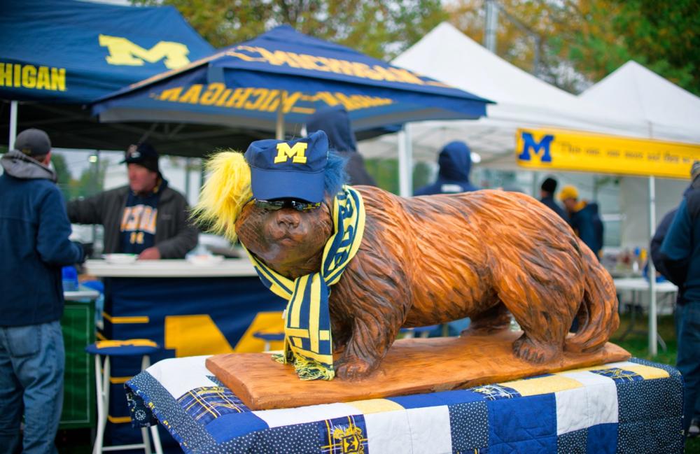 Wooden carving of wolverine wearing University of Michigan hat and scarf. Behind the wolverine are tents with M and Michigan printed on the tents.