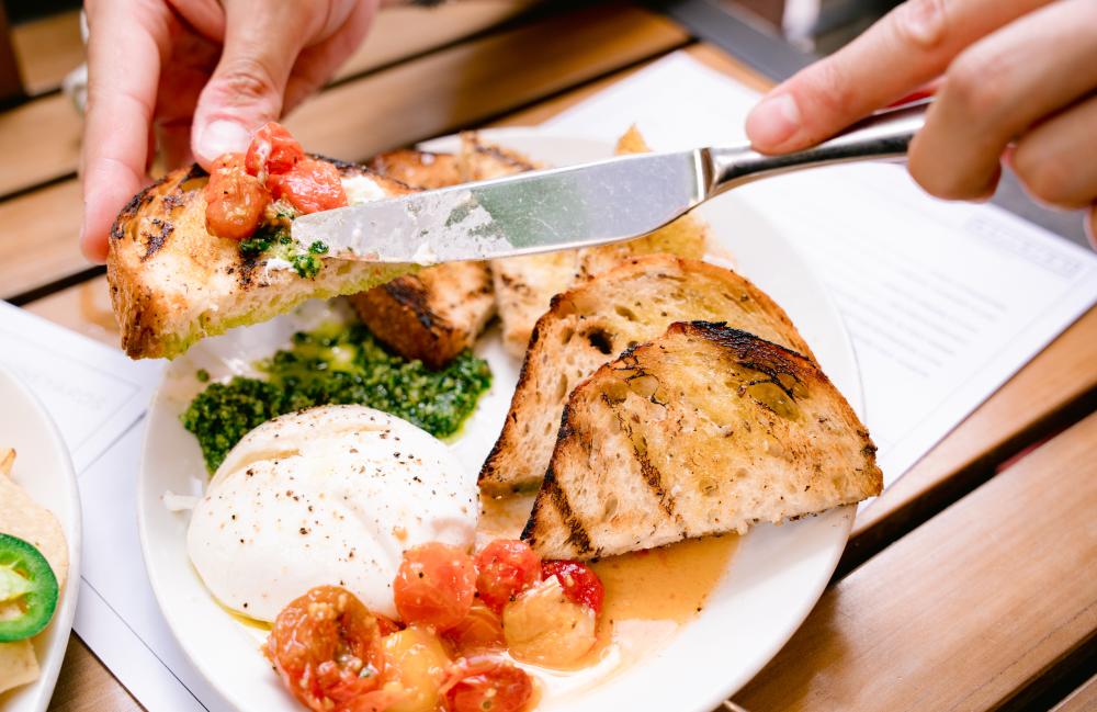 Person holding a piece of toasted bread, while using a butter knife to spread creamy burrata cheese and roasted tomatoes on the piece of toast.