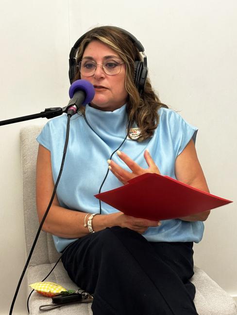 Marlo Christensen, Director of Marketing and Business Development for the Children's Museum of St. Tammany, speaks into a microphone during an All The Waves podcast recording. Wearing headphones and holding notes in a red folder, she gestures while discussing the museum's programs and mission.