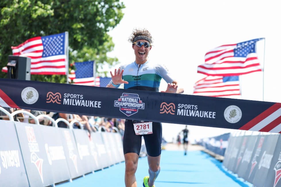 Triathlete crossing the finish line at the USA Triathlon National Championships in Milwaukee with American flags waving behind the race banner.