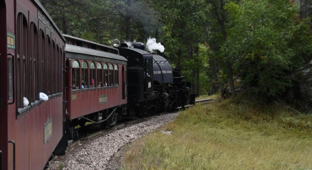 From the view of the passenger cars, a classic dark blue and black steam engine pulls dark red passenger cars around a curve along the Black Hills Central Railroad.
