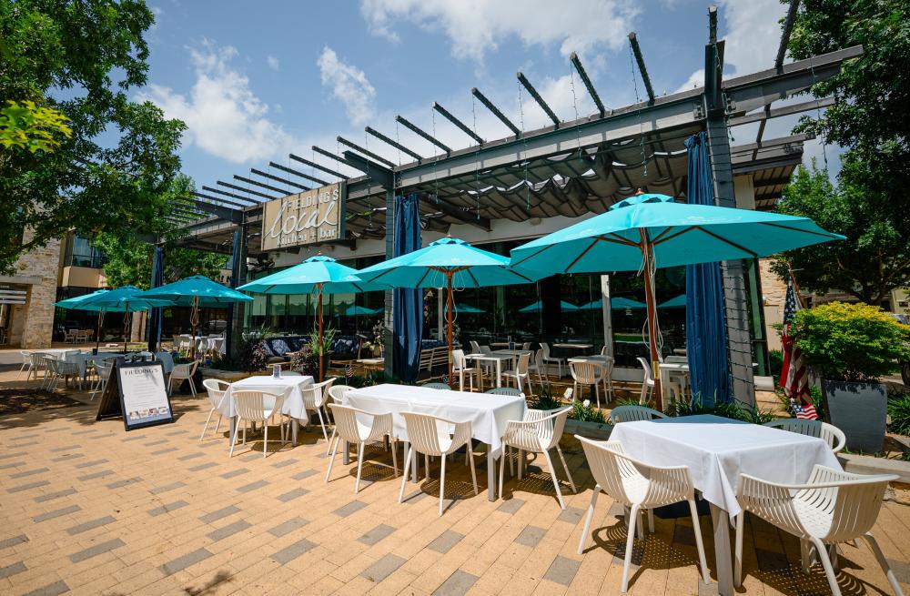 The entrance to Fielding's Local. The building is mostly glass with a shaded patio roof and a sign that reads, "FIELDING'S local kitchen + bar" in various font types. Patio tables are each shaded by large turquoise umbrellas.