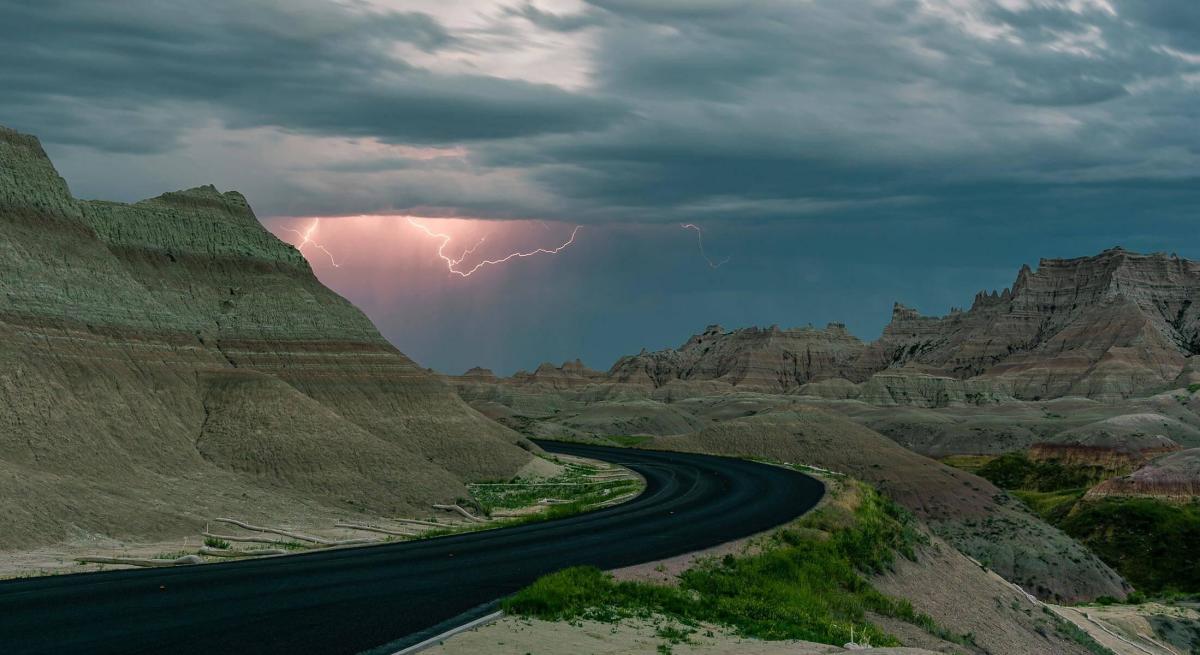 Road cutting through the badlands terrain with lightning striking in the distance