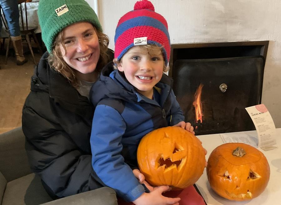 Child holding a carved pumpkin beside a wood-burning stove during a pumpkin decorating activity at Cleeve Common in the Cotswolds.