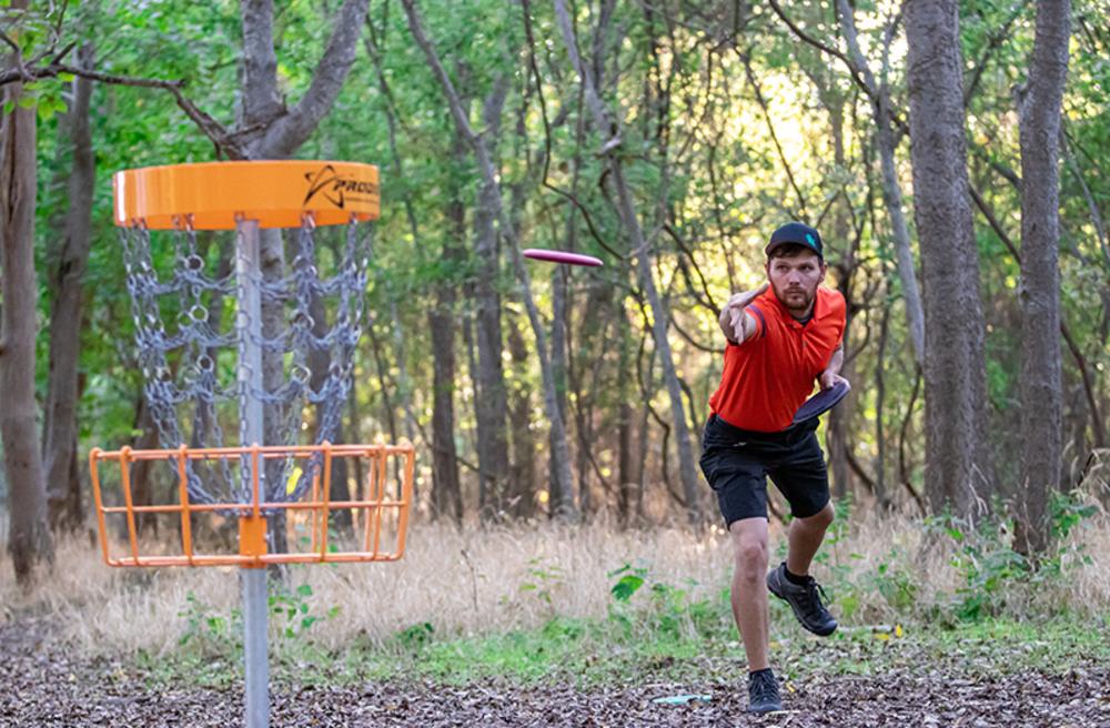 Man wearing a red shirt and black shorts tosses a frisbee towards an orange disc golf basket for the National Amateur Disc Golf Tour