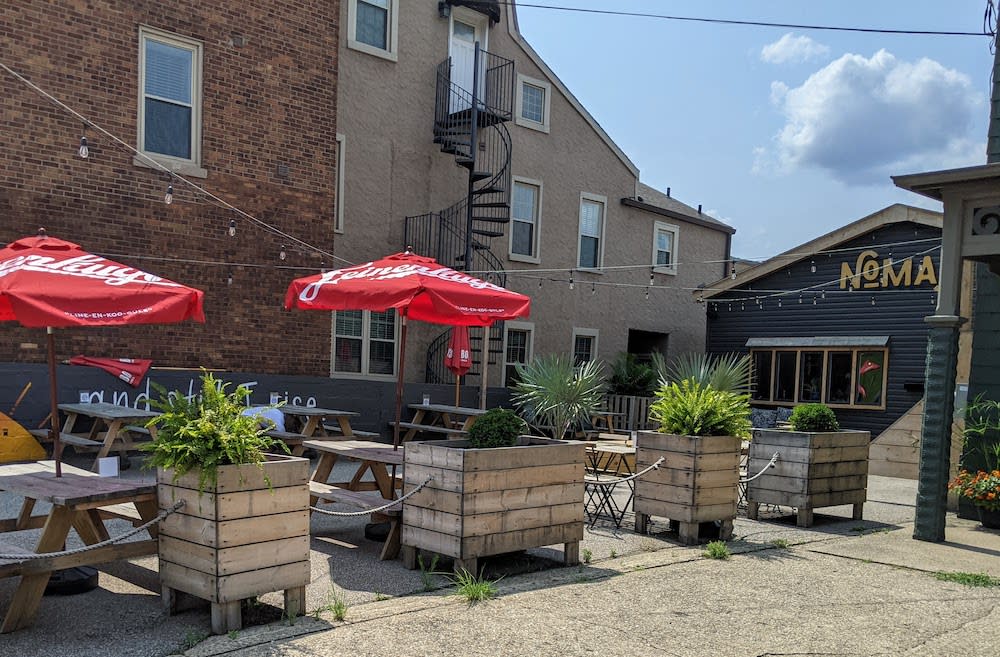 Multiple wooden picnic tables sit on a front patio outside of Nomad, a restaurant and bar with dark grey exterior