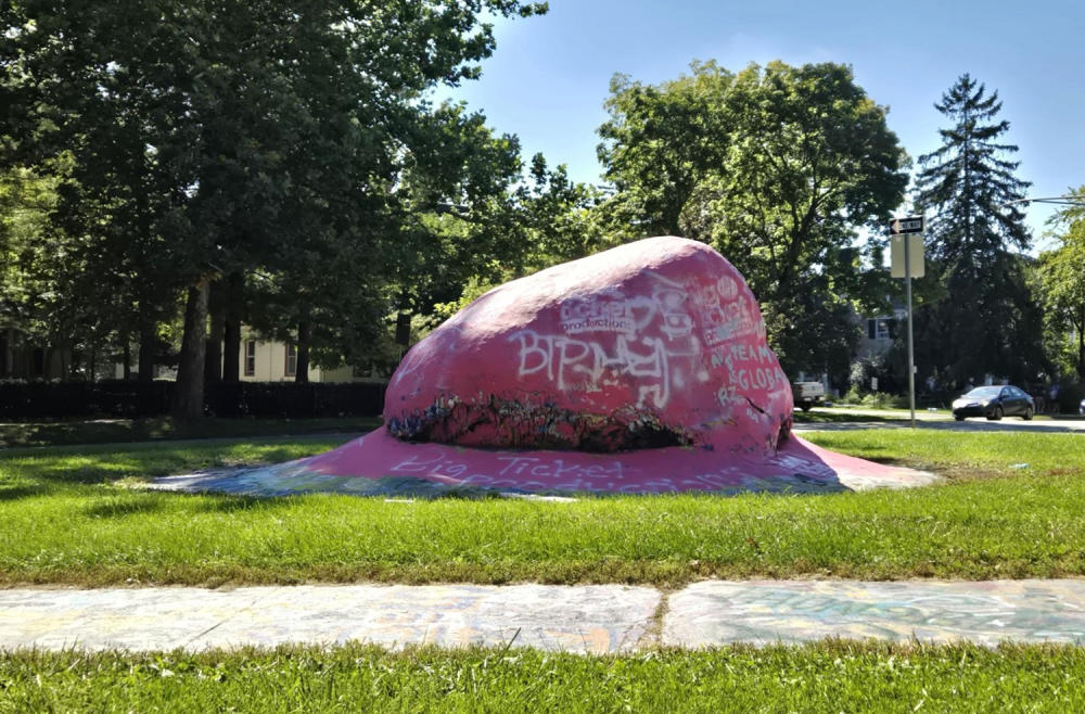 Giant rock at Washtenaw and Hill St painted pink with white spraypainted letters.  Greenery and trees surround the rock
