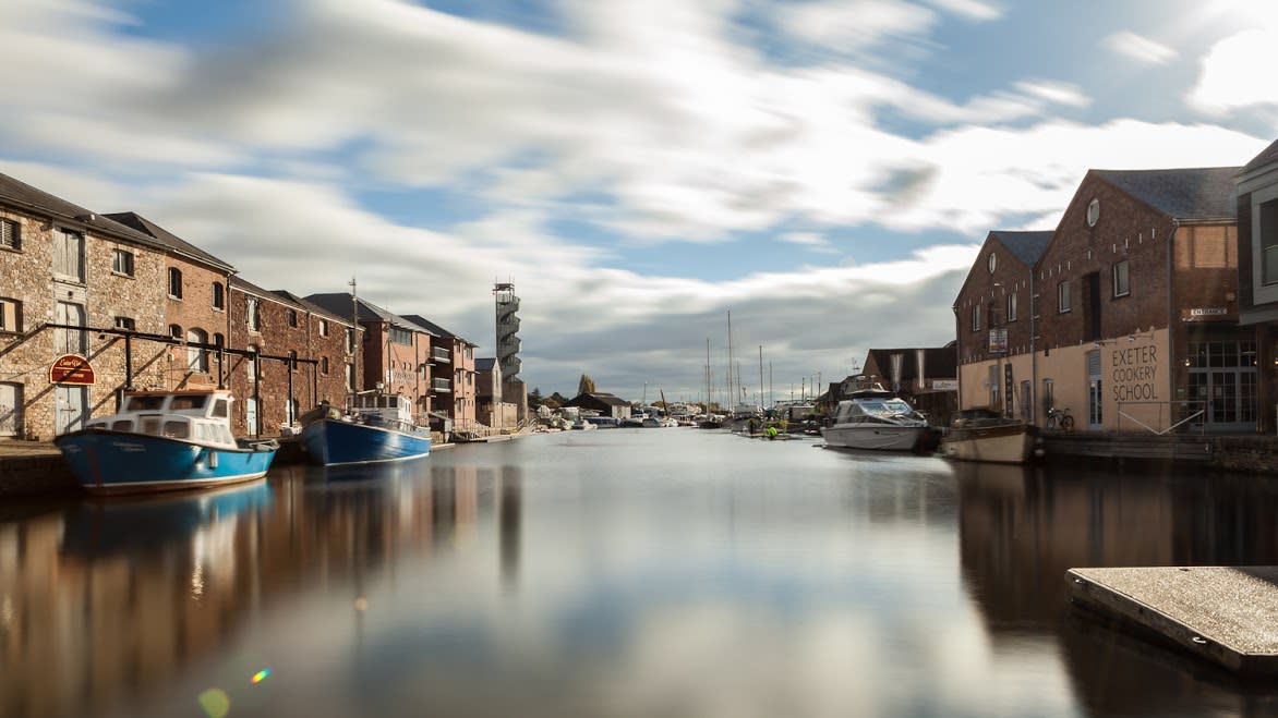 A serene waterfront scene with moored boats on a calm river, flanked by rustic brick buildings under a partly cloudy sky. A peaceful, charming atmosphere.
