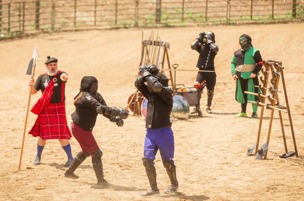Two people dressed in Celtic attire, fencing in an open sand arena outdoors. Two other people dressed in celtic attire watch as the fencers compete. A fith person coaches and points at the two competing.