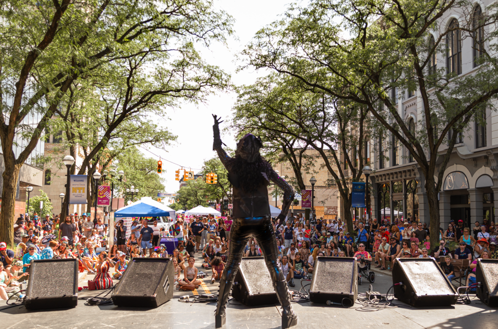 Person performs on a stage wearing all black. Their back faces the camera. The audicence watches, sitting and standing in the street with trees and buildings lining the left and right of the street.