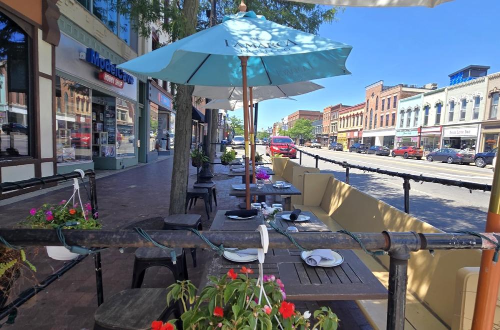 Patio table and chairs with umbrellas creating shade placed outside of C. King and Company, located on a sidewalk with a street and buildings lined up in the background.