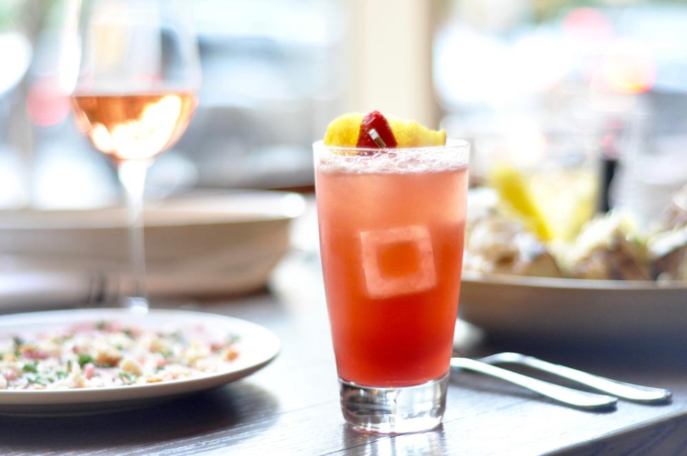 Pink cocktail with glass of wine and plates of food behind at Red Ash in Austin Texas