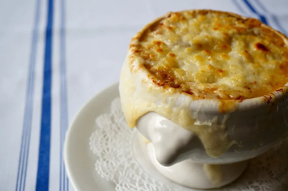 A steaming bowl of French onion soup topped with a thick layer of melted, golden cheese that’s bubbled over the edge of the crock, resting on a white plate with a lace doily beneath it.