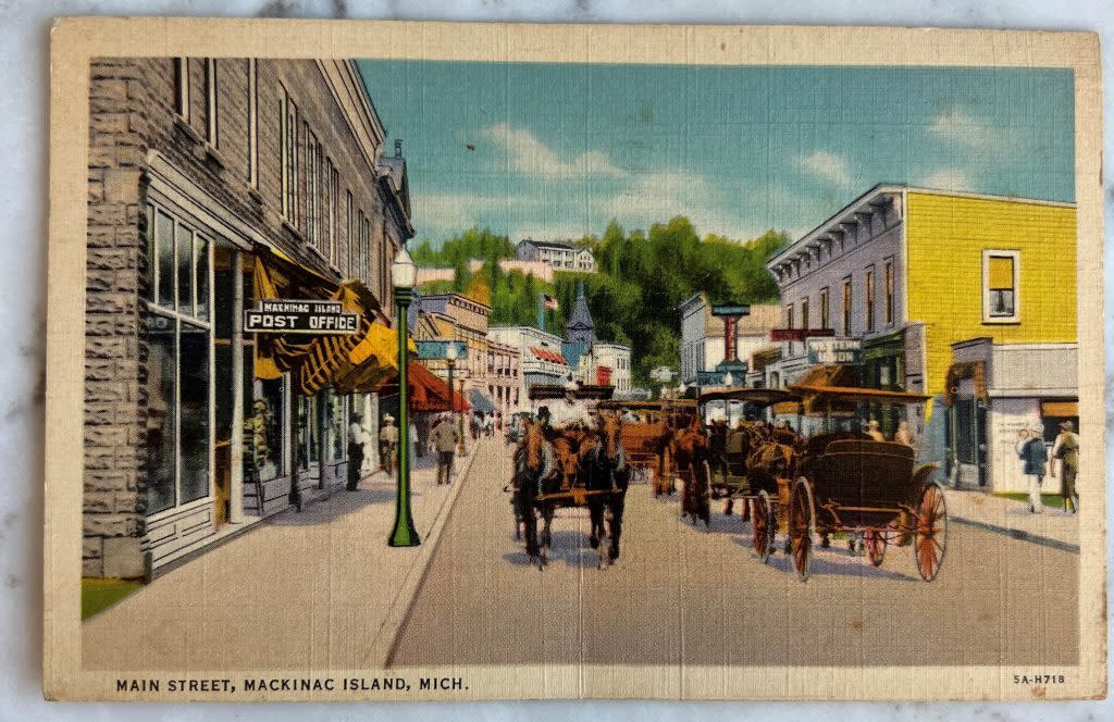 An old postcard showing horse-drawn carriages go past the Mackinac Island Post Office on Main Street
