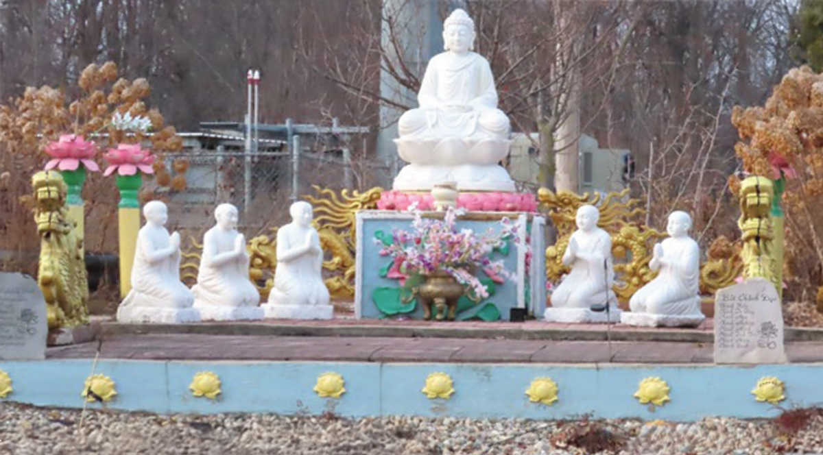 An outdoor Buddhist shrine shows a large central statue in a meditation pose with 3 monks kneeling on one side and 2 monks kneeling on the other. All cast in white rock.
