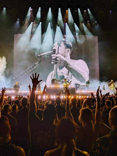 A packed crowd raises their hands at a live concert at The Orion Amphitheater, with a band performing on stage under bright lights and a large screen showing the lead singer mid-performance.