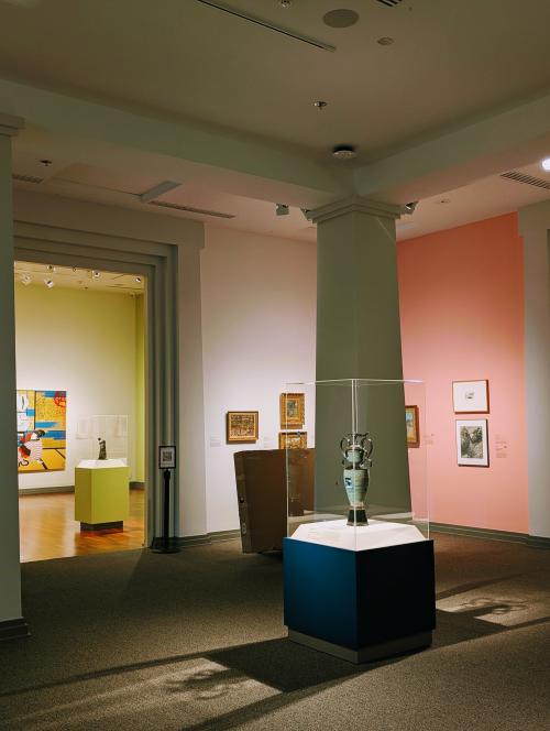 A gallery inside the Huntsville Museum of Art featuring a ceramic vessel displayed in a glass case, surrounded by framed artworks on colorful walls and softly lit exhibition spaces.