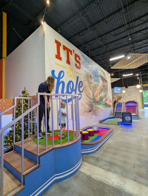 A child and adult play mini golf at Field Day, standing on a raised putting platform beside a colorful course. Bright signage, arcade-style games, and playful décor fill the indoor space, creating a fun, family-friendly atmosphere.