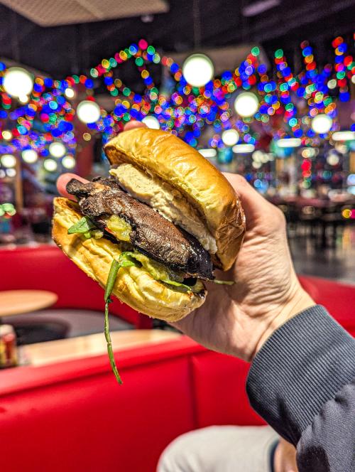 Hand holding a grilled mushroom sandwich with greens and a creamy spread inside Field Day, with colorful lights and red seating in the background.
