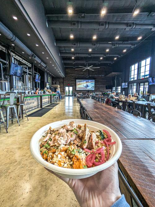 Hand holding a grain bowl topped with grilled chicken, roasted vegetables, pickled red onions and crumbled cheese inside Back Forty Beer Company.