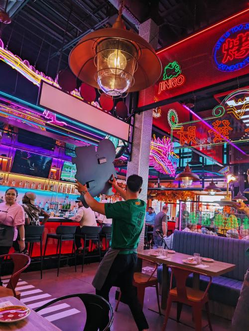 A server is holding up a large sign while walking through Dragon Alley restaurant in MidCity, surrounded by vibrant neon lights, lantern-style fixtures, and colorful signage. Guests sit at tables and along the bar, adding to the lively, high-energy atmosphere of the space.