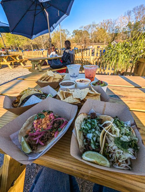 Street-style tacos served in trays with chips, dips, and margaritas on a wooden picnic table at an outdoor patio. In the background, people sit under umbrellas near a fence and greenery, creating a casual, sunny dining atmosphere.