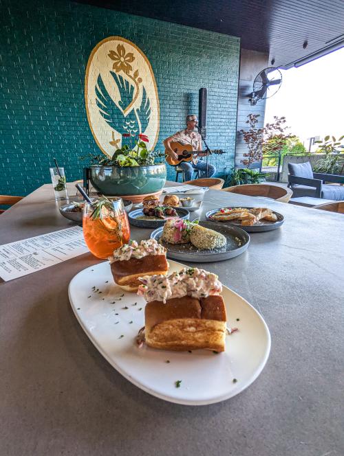 Plate of slider sandwiches topped with a creamy salad sits in the foreground on a restaurant table, with cocktails and shareable dishes nearby. In the background, a musician plays guitar on a patio with greenery, seating, and a teal mural wall, creating a relaxed live music dining atmosphere.