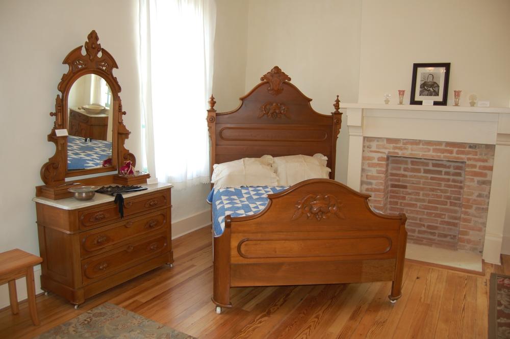 An antique wooden bed with blue and white quilt next to a wooden dresser with mirror in the bedroom in the Susanna Dickinson House museum in austin texas