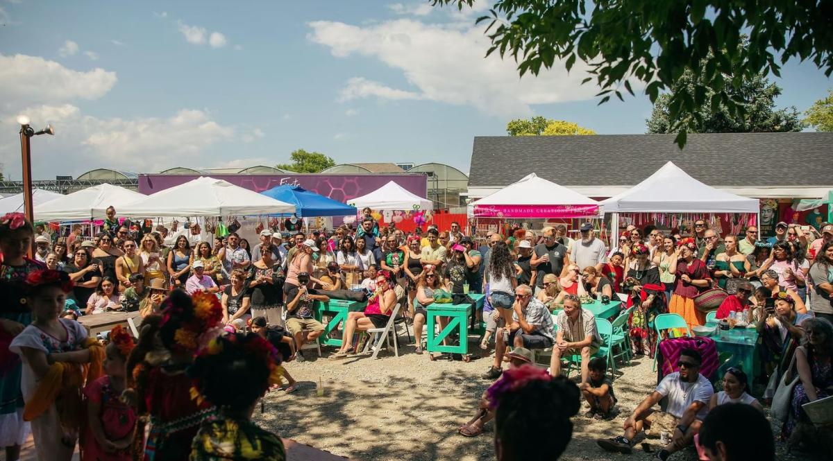 Crowd gathered at an outdoor cultural festival watching children in traditional Mexican clothing perform, with vendor tents and colorful decorations in the background under a sunny sky.