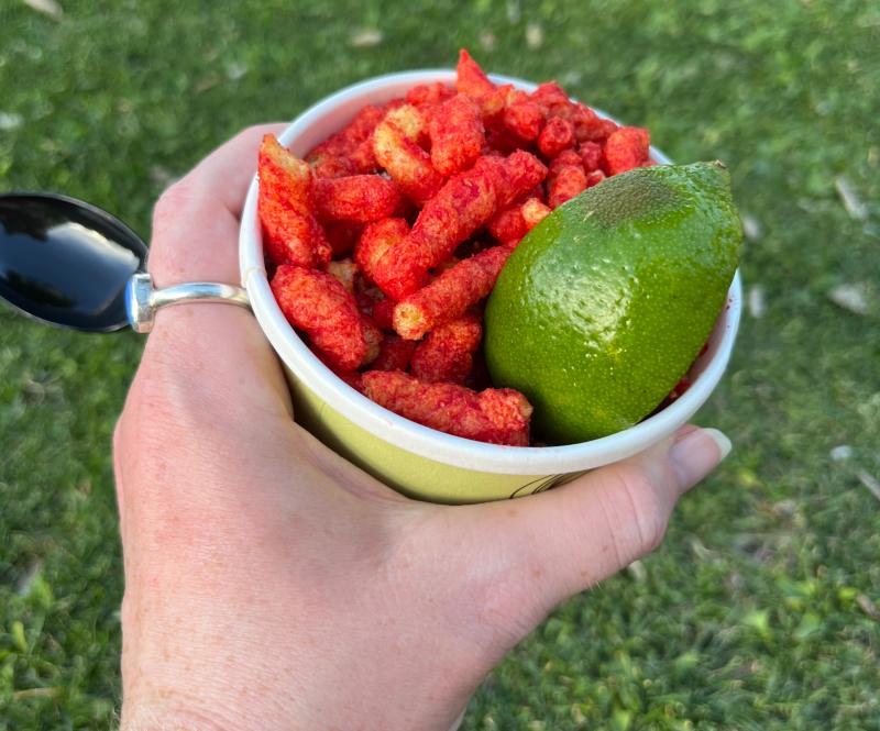 Someone holds a cup of hot elote covered in Hot Cheetos with a lime wedge.